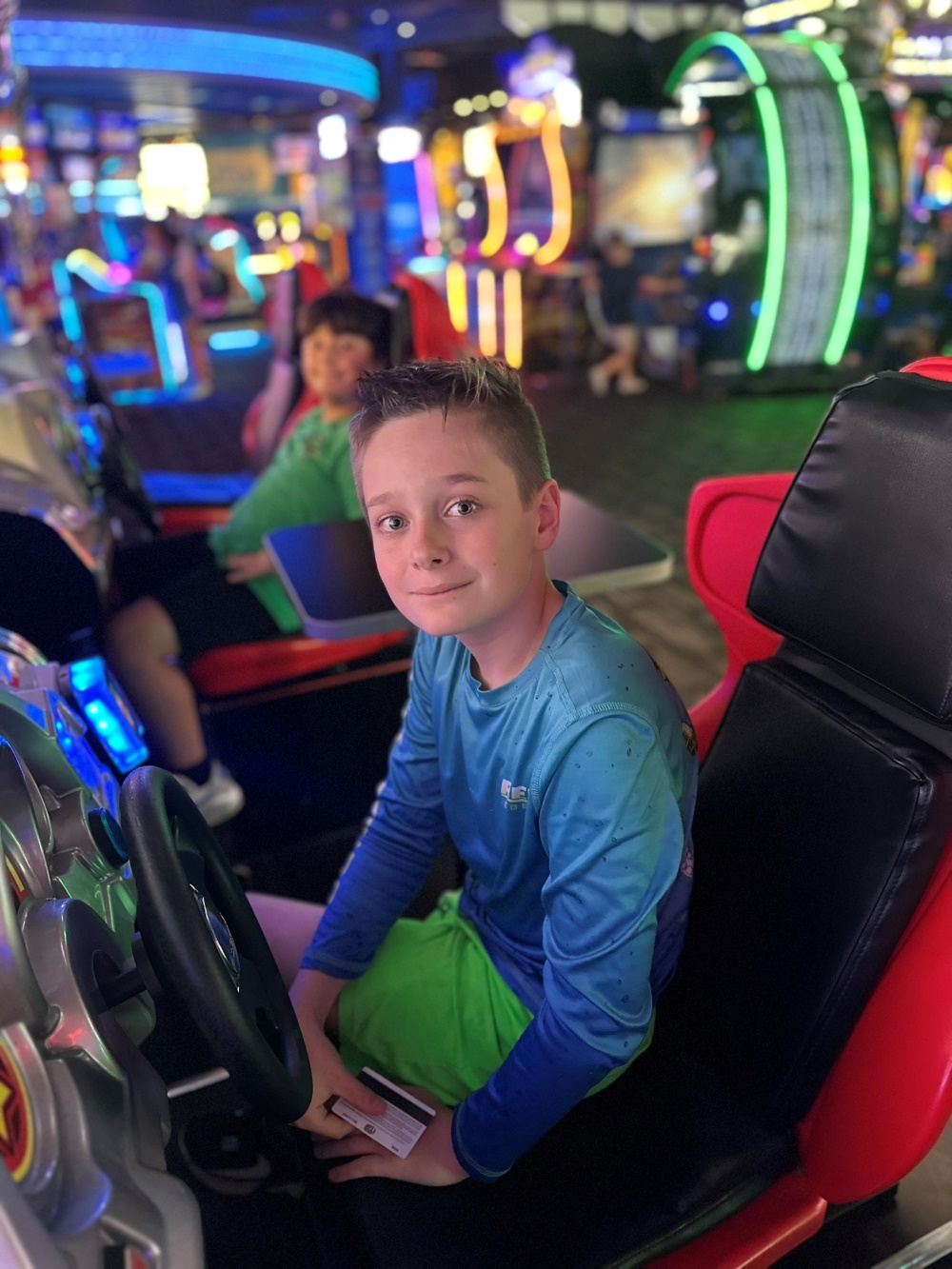 Boy sits in an arcade racing game seat holding a play card, with colorful machines in the background